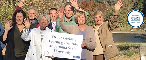 A group of seniors standing in front  of a lake on the Sonoma State campus holding an Osher Lifelong Learning sign