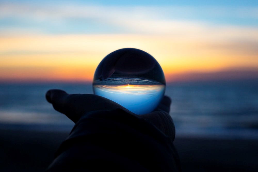 A hand holding up a clear glass globe against the backdrop of the ocean