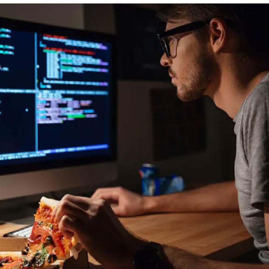 A man looking at computer code on a monitor while picking up a pizza slice